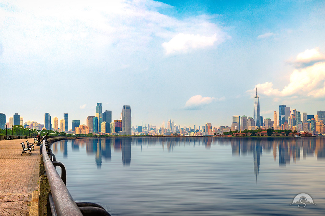 From a quiet waterfront promenade lined with empty benches, this serene photograph captures the Lower Manhattan and Jersey City skylines reflected in calm harbor waters beneath a soft, cloud-filled sky.