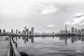 From a quiet waterfront promenade lined with empty benches, this serene black and white photograph captures the Lower Manhattan and Jersey City skylines reflected in calm harbor waters beneath a soft, cloud-filled sky.