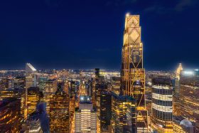 A gleaming, lattice-crowned skyscraper rises above Manhattan's glittering nighttime skyline, its golden facade illuminated against the deep blue night.