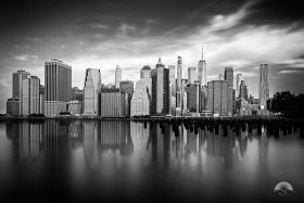 A serene black and white long-exposure captures Lower Manhattan's skyline mirrored perfectly in the still waters of the East River, with weathered pier pilings and dramatic streaked clouds adding timeless atmospheric depth.