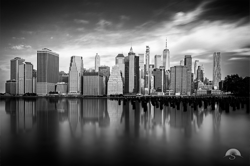 A serene black and white long-exposure captures Lower Manhattan's skyline mirrored perfectly in the still waters of the East River, with weathered pier pilings and dramatic streaked clouds adding timeless atmospheric depth.