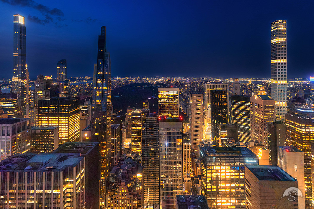 Slender supertall towers glow gold against a deepening blue twilight sky, encircling the dark, tranquil void of Central Park in this striking aerial view of Manhattan's ever-evolving skyline.